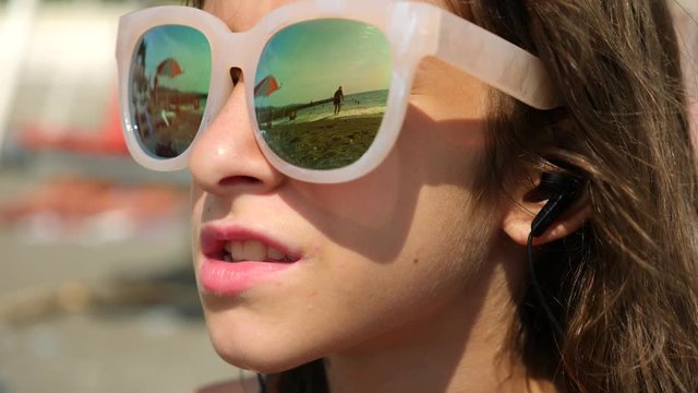 Teenage girl in sunglasses. Sits on the beach and sings a song listening to her in headphones with a smartphone. In the glasses reflects the beach with sea waves. 4k. Slow motion