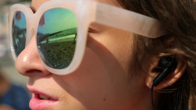 Teenage girl in sunglasses. Sits on the beach and sings a song listening to her in headphones with a smartphone. In the glasses reflects the beach with sea waves. 4k. Slow motion