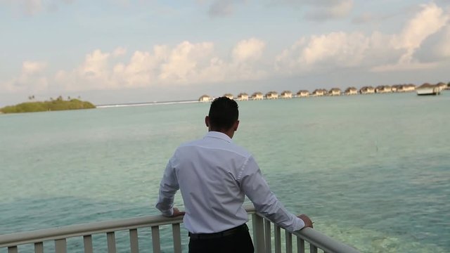 One businessman in official suit standing on the balcony of his villa bungalow and looking to the beautifull blue lagoon at luxury tropical hotel resort. Rich wealth lifestyle concept