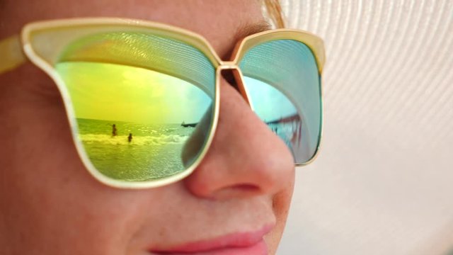 Close-up face of a happy woman in sunglasses. The beach with the resting people is reflected in the glasses. 4k, slow motion