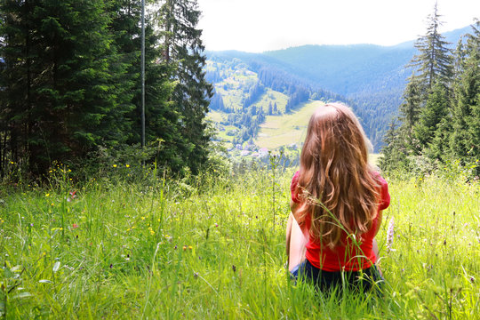 Young brown-haired woman female with curly hair sitting at green grass glade at mountain landscape background in sunny summer day. Carpathian Mountains, wild mountain landscape Ukraine, Vorohta