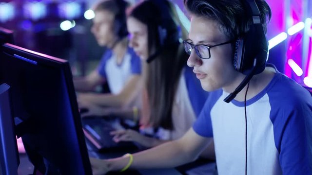 Professional Boy Gamer Plays in Video Game on a eSports Tournament/ Internet Cafe. He Wears Glasses and Headphones and Speaks into Microphone. Other Girls and Boys Players Playing in Background. 