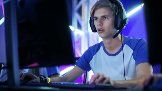 Professional Boy Gamer Plays in Video Game on a eSports Tournament/ in Internet Cafe. He Wears  Headphones and Speaks Commands into Microphone.  Shot on RED EPIC-W 8K Helium Cinema Camera.