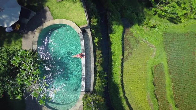 AERIAL TOP DOWN Happy young woman enjoying summer vacation in luxury tourist resort above gorgeous rice terraces in sunny Bali. Girl in infinity pool above lush palm tree jungle on tropical island
