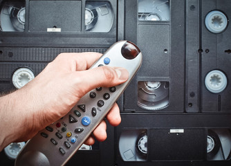 Close-up TV remote control in hand on a background of video cassettes. Retro media technologies. Entertainment 90s.