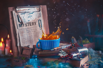Tea splash in a ceramic cup on a wooden background with candles, mystery newspaper clips, books, leaves and moss
