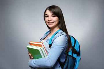 Young smiling girl with knapsack and books on grey background