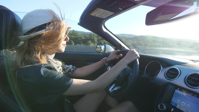Blond model girl is driving in convertible car in white hat in California having her hair waving in the wind
