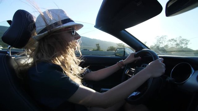 Blond model girl is driving in convertible car in white hat in California having her hair waving in the wind