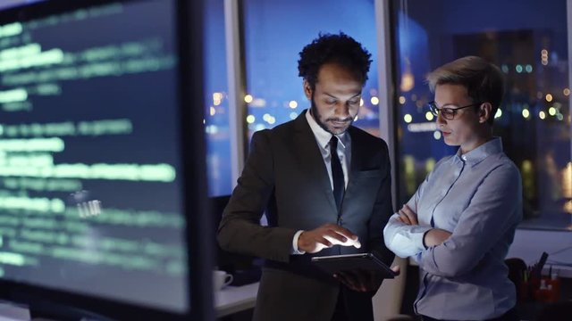 Tracking shot from computer monitors with programming codes to female IT professional discussing something on tablet with male African colleague in the office at night