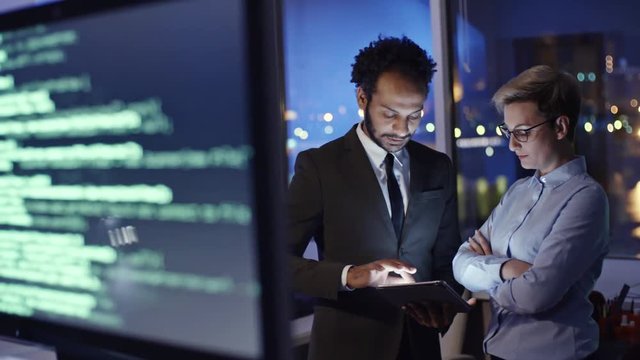 Tracking shot of female cyber security analyst discussing something on tablet with male African colleague in the office at night and computer monitors with programming codes