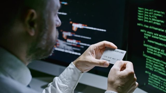 Closeup of businessman sitting at desk in front of computer screens with program codes, reading number on credit card, putting it on the table and typing on keyboard