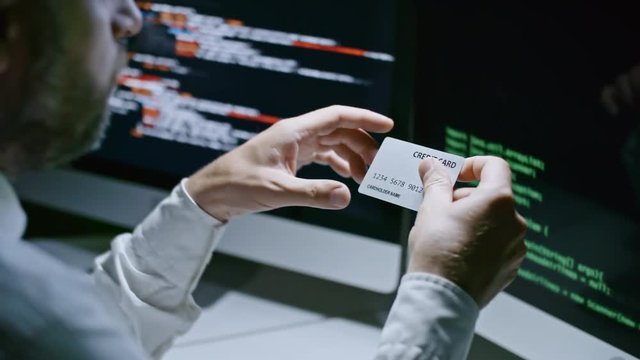 Closeup of businessman sitting at desk in front of computer screens with program codes and reading number on credit card