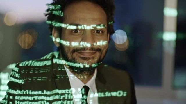 Tilt up of African programmer typing code on computer in the dark office, looking at camera and smiling. Green source code reflecting on man