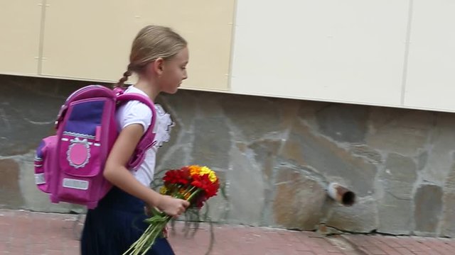 Sorrowful girl go back to school with backpack and bouquet flowers. Young white caucasian girl with pink baclpack walking along stone wall of school. Handheld shot walking serious elementary girl.