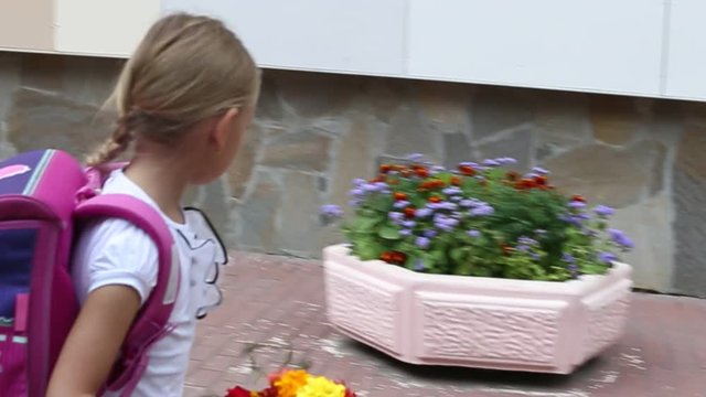 Sorrowful girl go back to school with backpack and bouquet flowers. Girl fright some thing at flowerbed. Young white caucasian girl with pink baclpack walking along stone wall of school. Handheld shot