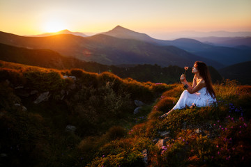 A girl sits on the edge of the cliff and looking at the sun valley and mountains. Woman sitting on mountain top and contemplating the sunset. Young woman in long white dress - 168407536