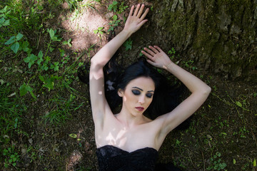     Pale woman in black dress lying on the ground in woods, dark mystery scene 