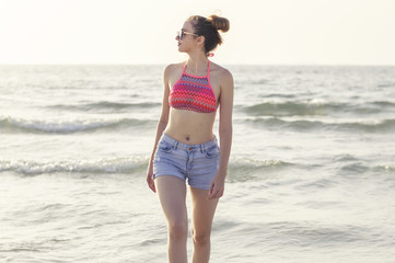 Fototapeta na wymiar Young woman in blue denim shorts and colorful swimsuit top at the beach. Young female walking at the seashore. Vacation, freedom and enjoyment