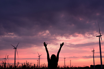 Fototapeta na wymiar silhouette of woman with rised up hands and electric windmills in sunrise sky, alternative energy concept