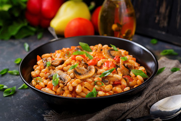 Stewed white beans with mushrooms and tomatoes with spicy sauce in a black bowl.