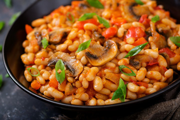 Stewed white beans with mushrooms and tomatoes with spicy sauce in a black bowl.