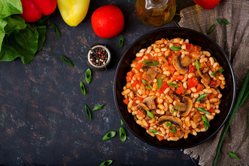 Stewed white beans with mushrooms and tomatoes with spicy sauce in a black bowl. Top view. Flat lay