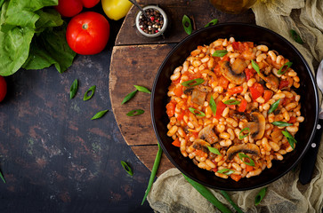 Stewed white beans with mushrooms and tomatoes with spicy sauce in a black bowl. Top view. Flat lay