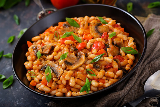 Stewed white beans with mushrooms and tomatoes with spicy sauce in a black bowl.