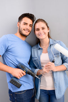 studio portrait of a young couple on grey plain background holding cordless drill do it yourself tool and white paint brush