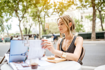 Young stylish woman reading journal sitting outdoors at the cafe in Paris