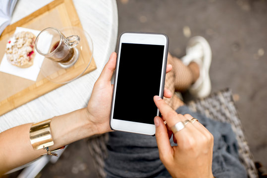Woman holding a smart phone with empty screen sitting outdoors at the cafe with cake and coffee on the table