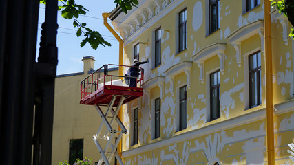 A few workers in blue uniform are restoring a yellow facade of a building from red crane lifter.