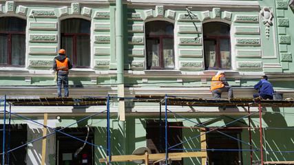 Three men are restoring a birch facade of an old building. Men in red color clothes are standing on scaffolding.