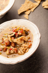 
White chicken chili with red peppers, chicken, and beans in white bowl with corn tortilla chips angled shot

