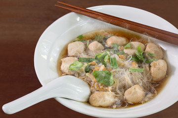 White noodle with pork ball with herb Chinese soup in white bowl with chopsticks and spoon on brown wooden fbackground  thai food