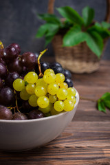 Close up bowl of various grapes: red, white and black berries on the dark wooden table with wicker basket with leaves in the background . Selective focus.