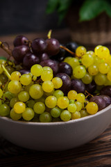 Close up bowl of various grapes: red, white and black berries on the dark wooden table with wicker basket with leaves in the background . Selective focus.