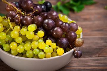 Close up bowl of various grapes: red, white and black berries on the dark wooden table. Selective focus.