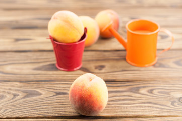 One fresh whole ripe peach in red metal bucket and three peaches separately and orange watering can on rustic old wooden planks