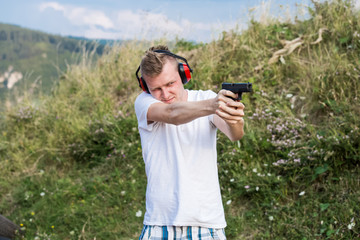 Young handsome blonde guy training policeman special forces shooting with the  pistol gun aiming at target with hands and protective gear in the nature in sunny day