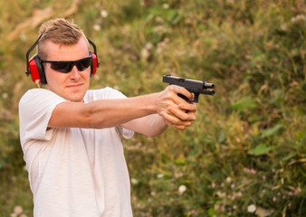 Young blonde handsome guy shooting from fire arms gun violent  pistol aiming at the enemy target with firing bullets with black police sunglasses and sound blocker