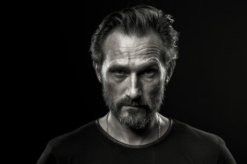 Black and white photo of mid aged man showing severe emotion. Beardy male in black t-shirt on dark background with serious look.