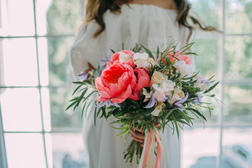 Bride holds in hands a rustic wedding bouquet with white roses and crimson peonies on window background. Close-up
