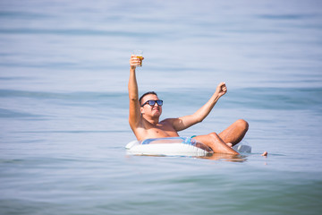 Young man with hands up in sunglasses with a glass of beer floating on rubber ring in the ocean water at sunset