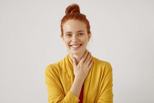 Headshot of cute redhead girl with hair bun wearing yellow cardigan looking at camera with shy charming smile and holding her neck, touched by nice compliment. Positive human emotions and feelings