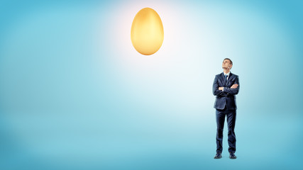 A businessman with crossed hands looks up to a large shining golden egg overhead.