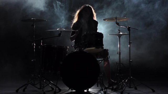 Girl is kicks from playing drums, playing energetic music. Black smoke background. Silhouette. Slow motion