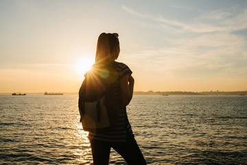 A young tourist girl with a backpack stands next to the sea at sunset and looks into the distance. Rest, relaxation, travel, vacation.