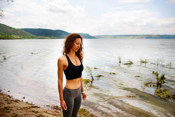 A beautiful sporty woman standing on the shore of a lake in sportswear and preparing for an exercising